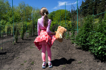 A 7 year old girl with dark hair in a pink suit stands alone in a green garden on the ground with a...