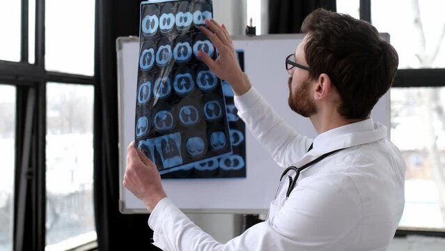 Attractive smart doctor looking at x-ray at his cabinet, caucasian male therapist in white medical uniform and glasses concentrated learning patient's x-ray, standing in his medical office
