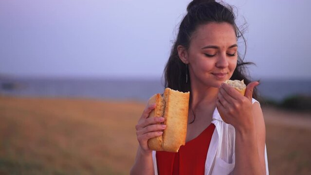 Medium shot smiling young Caucasian woman smelling delicious organic bread sitting on the right at background of yellow field and blue sea. Portrait of happy farmer with pastry outdoors