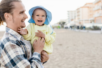 Fototapeta premium Positive man holding baby girl in panama hat on beach in Treviso.