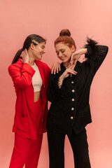 Positive young caucasian girls with smile on face and closed eyes pose on pink background. Brunette, redhead wear trendy shirts, pants. Lifestyle, female beauty concept