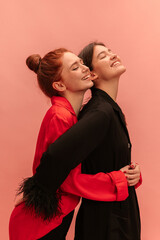 Joyful young сaucasian women close eyes and smile with heads up against pink background. Models brunette and redhead wearing shirts embrace each other. Lifestyle, different emotions, leisure concept. 