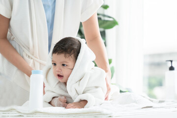 Cute Caucasian little toddler baby girl wear bathrobe after bathing is smiling and lying on towel while mother wipe and apply talcum powder on her body. Hygiene care for children concept - obrazy, fototapety, plakaty