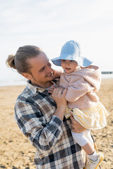Young long haired dad holding toddler child on beach in Treviso.
