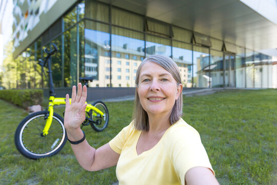 Active cheerful senior woman looking at monitor screen taking selfie camera, talking on video call, outside near modern office building, cycling