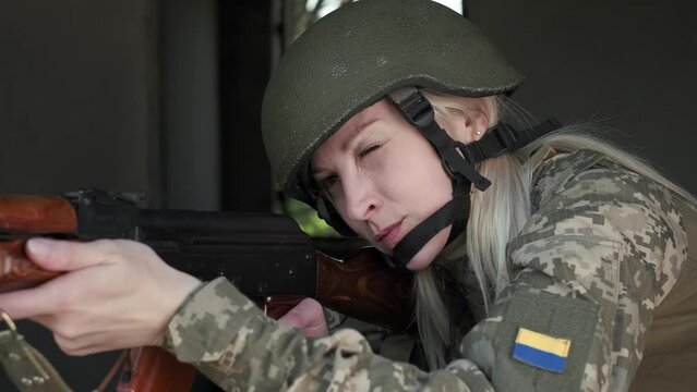Close-up armed young woman in camouflage uniform in zone of armed conflict aims with an assault rifle from window of destroyed building, slow motion. Russian-Ukrainian war