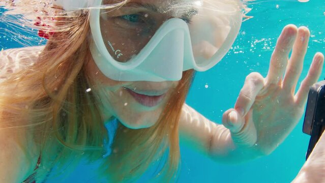 Young woman swims underwater with action camera and shows OK signal with her hand