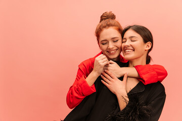 Cute young caucasian girls with their eyes closed are standing in embrace on pink background with place for text. Brunette and redhead are wearing black and red shirt. Different emotions concept. 