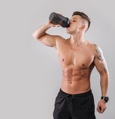 Naklejka premium Athletic muscular handsome guy with a naked sporty body with a tattoo drinking water from a black bottle against a gray background. Healthy lifestyle and diet