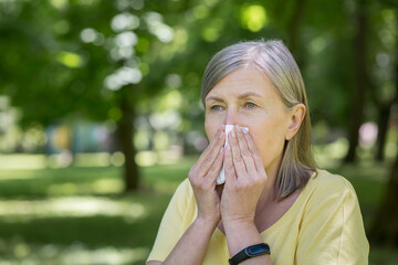 An elderly woman in the park with allergies has a runny nose and sneezes near trees - obrazy, fototapety, plakaty