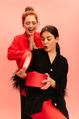 Two young caucasian girls with positive emotions look at gift on pink background. Brunette looks...
