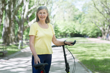 Fototapeta premium Portrait of senior active woman, gray-haired woman in park looking at camera and smiling with bicycle, summer day