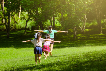 Naklejka na ściany i meble Two lovely daughters running with their mother spreading their arms as if flying in a spring garden. Rear view of young mother playing with children in the park. Happy family having fun in the park.