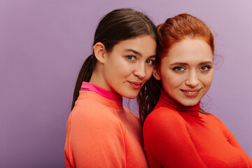Close-up of young caucasian woman looking confidently at camera against purple background. Brunette and redhead wear bright sweaters. Lifestyle, different emotions, leisure concept. 