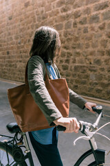 Fototapeta na wymiar A woman with her black urban bike, looking at an old stone wall, in the city of Barcelona. A sunlight reflection on her hair. She is wearing a blue shirt, a gray jacket and a large tan bag. 