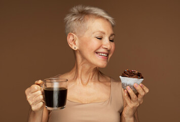 smiling senior woman with mug of coffee and chocolate cupcake over brown background.