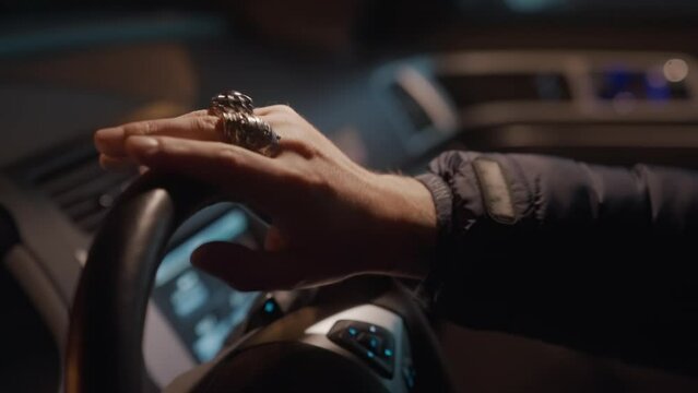 At night in the parking lot, a lonely man in a jacket in the front seat of the car impatiently taps the steering wheel with his fingers with metal skull rings on, slow motion close-up.