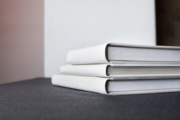 Three white stylish large photobooks with a leather cover lie on a dark gray surface against a light wall in the room.