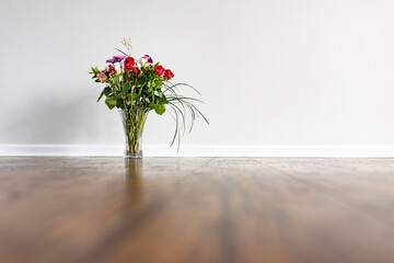 A vase with a rose bouquet placed on hardwood floor next to a wall with baseboard in an empty...