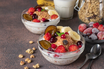 Granola crispy muesli with natural yogurt, frozen berries and fruit and nuts in two glass bowls on brown background, healthy breakfast