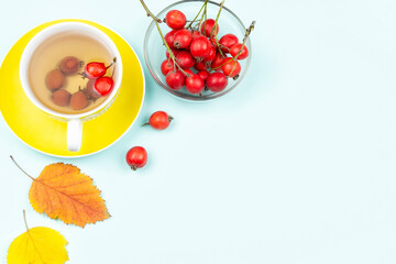 Autumn concept. A bunch of fresh hawthorn berries in a plate, a Cup of tea (infusion) of hawthorn on a saucer and yellow leaves on a blue background. Space for text.