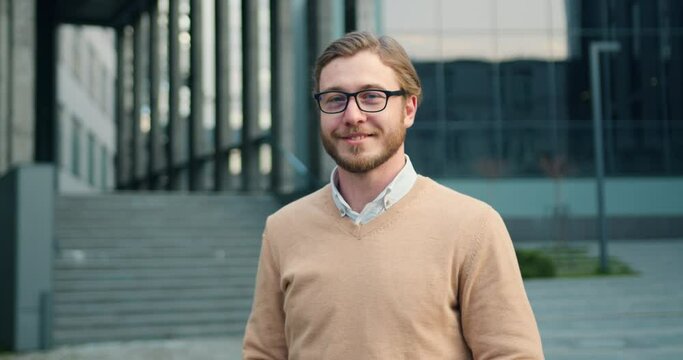 Bearded young business man in glasses smiling on camera while standing outdoors by modern office building. Portrait of positive male person dressed in stylish formal clothes