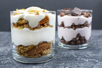 Cottage cheese dessert with oatmeal cookies and yogurt in a glass glass on a black (dark) background. Chocolate dessert in the background.