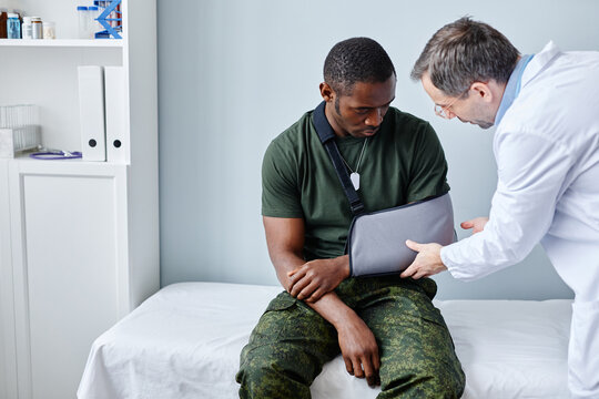 Mature Caucasian traumatologist palpating young African American soldiers broken arm in sling during appointment