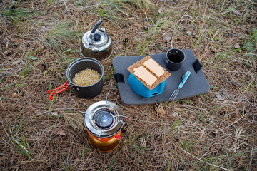 Camping meal breakfast on the grass, cheese sandwiches, noodle pot, gas burner, metal kettle with...