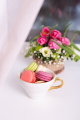 
Sweet dessert, macaroons in golden mug and pink tulips on window background

