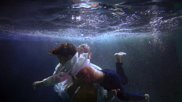 a boy in a white shirt and jeans is swimming underwater. father supports him