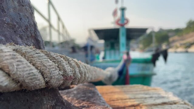 Rusty mooring bollard old cast iron close up with rope tie docking fishing boat floating at pier. Boats floating on shore water sunny day. Flag on boat flapping windy day