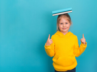 Little girl in yellow hoodie having fun holding books on her head on blue background with copy...