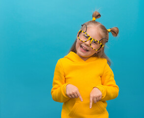 Naklejka premium Funny playfull little girl in bee glasses and yellow hoodie on blue background pointing down to free copy space.
