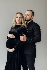 Husband and pregnant blonde wife in a black dress in the studio on a black background stand and hug