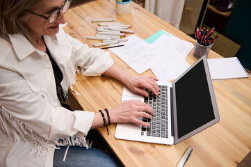 Top view of a pleasant senior woman dressmaker typing text on laptop keyboard sitting at a wooden desk in fashion design atelier. Tailoring scissors and fashion sketches on the table