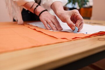 Fototapeta premium Tailor at work. Close-up of a dressmaker's hands transferring sewing patterns for clothes on orange colored satin fabric in fashion design atelier