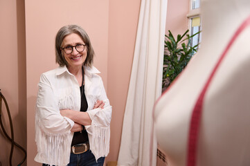 Elegant mature European woman dressmaker, fashion designer, smiling, standing with her arms crossed next to a mannequin in her own fashion atelier