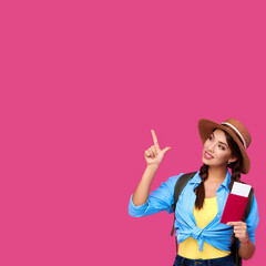Smiling girl student holding passport with airplane ticket and showing product via finger gesture on pink isolated background. Female traveler wearing straw hat. Woman student with backpack