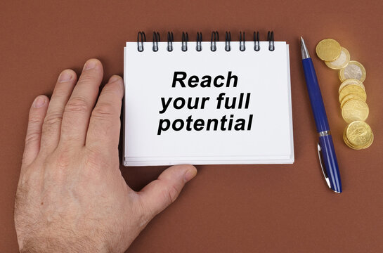 On A Brown Surface Are Coins, A Pen And A Notepad With The Inscription - Reach Your Full Potential