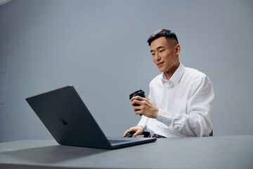 Asian man in white shirts glass of coffee sits in front of a laptop office Gray background