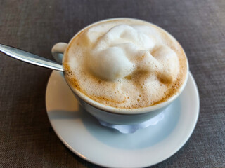 View of a mug of a cappuccino coffee with fluffy, white milk froth on the table covered with neutral grey tablecloth in front of window. Hot drink in a Café