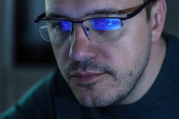 Portrait of millennial concentrated trader man in glasses looks at screen with graphs in dark. Caucasian man monitors exchange rate. Business Graph and chart of currency reflection in eyeglasses.