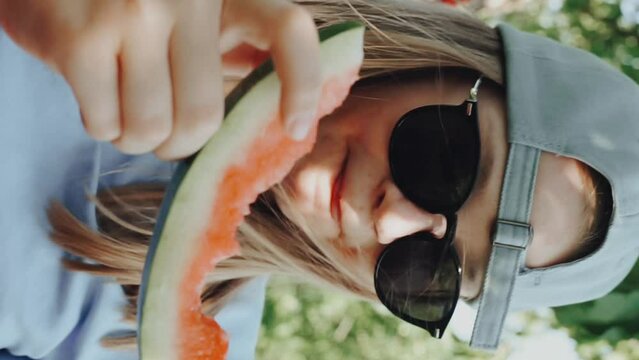 Candid portrait hipster Tin sunglasses een eating watermelon in backyard garden in hammock. Happy Kid eat fruit outdoors. Healthy snack for children. Summertime girl biting juicy slice of water melon.