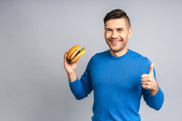 Young ukrainian man holding a piece of hamburger. Student eats fast food. Burger is not helpful food. Very hungry guy. Diet concept. Isolated over grey white background. Thumbs up.