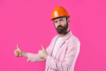Portrait of young construction engineer wear orange hard hat, in a pink jacket standing on red studio background. The man points with his hand.