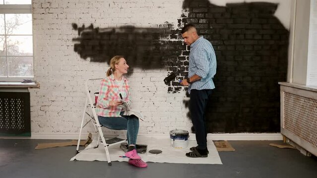Caucasian young and cheerful man and woman talking and deciding how to decorate a room and colour of walls while having repair at home and sitting on the floor ladder...