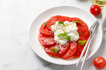 Fresh salad with delicious sliced tomatoes and Stracciatella cheese on white plate with fork, oil,...