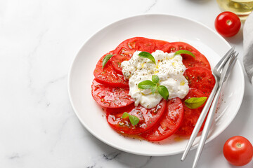 Fresh salad with delicious sliced tomatoes and Stracciatella cheese on white plate with fork, oil, basil and tomatoes. Copy space.