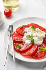 Salad with Stracciatella cheese on white plate with fork, oil, fresh basil and tomatoes. Italian Stretched curd cheese product in Stracciatella salad. Close up